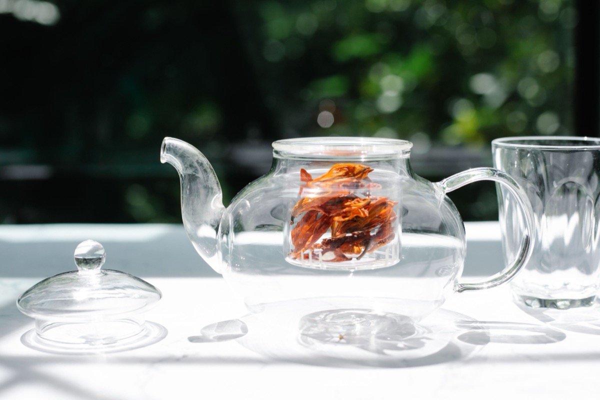 Théière en verre contenant des fleurs séchées en infusion, posée près d'un verre dans la lumière naturelle