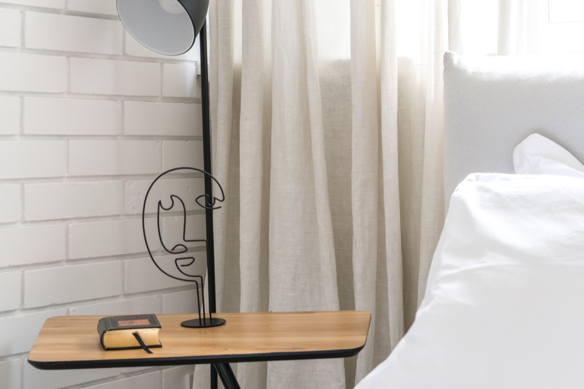 Table de chevet en bois avec un carnet et un stylo, lampe et rideau clair près d'un lit