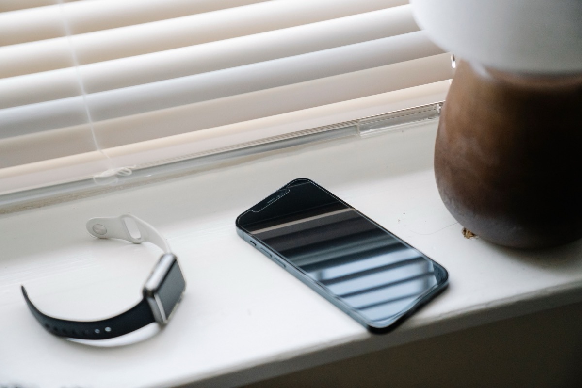 Montre connectée à écran éteint et téléphone posés sur un rebord de fenêtre, devant un store baissé