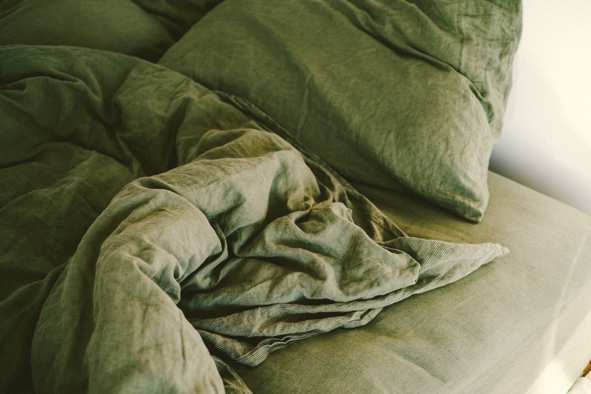 Couette et drap en lin vert froissés sur un lit défait, en lumière naturelle