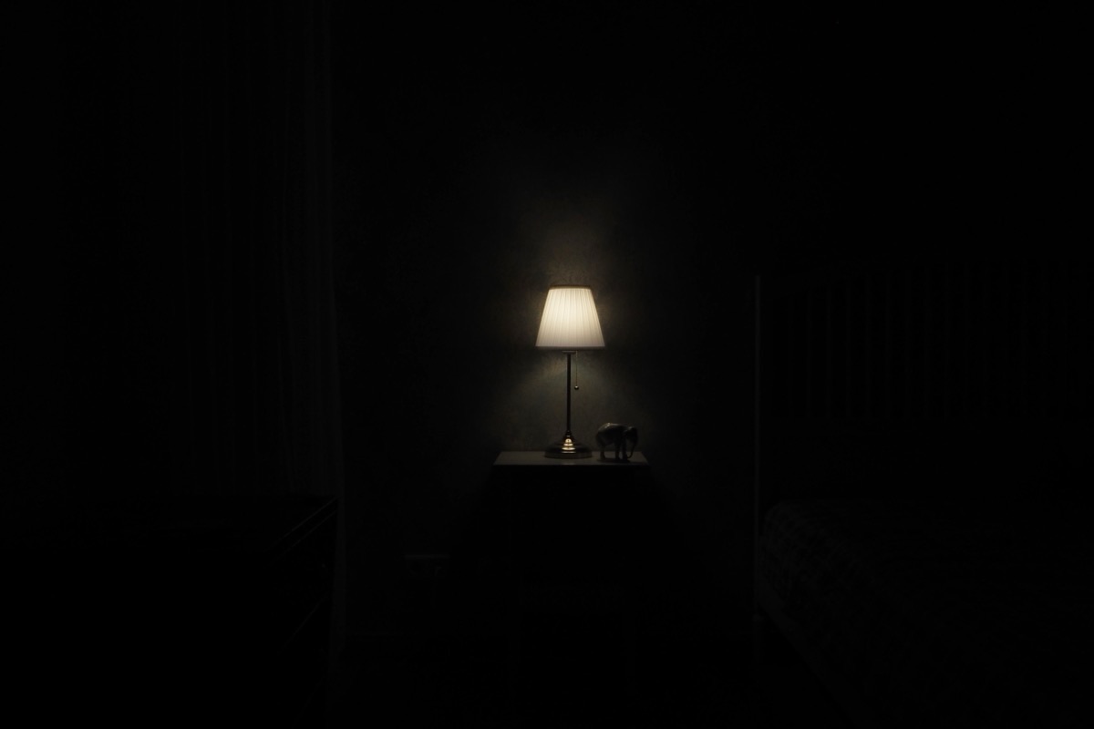 Lampe de chevet allumée diffusant une lumière douce dans une chambre plongée dans l'obscurité, bord du lit visible à droite
