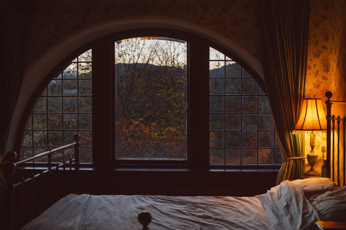 Chambre ancienne avant le lever du jour, lit défait au premier plan devant une grande fenêtre cintrée et une lampe de chevet allumée