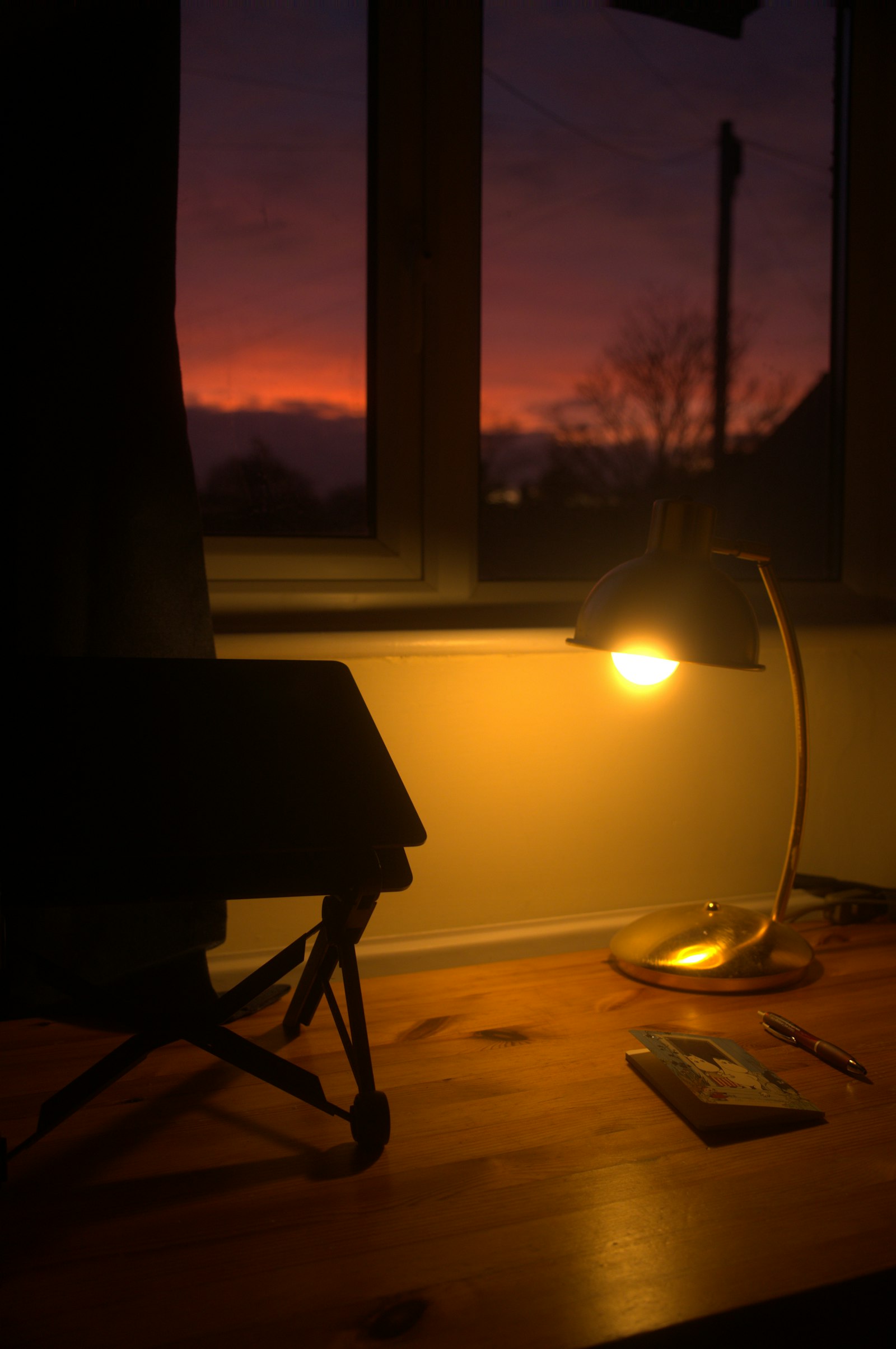 Bureau avec lampe allumee et coucher de soleil par la fenetre, fin de journee de teletravail